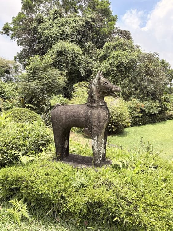 A weathered stone horse sculpture standing on a clipped lawn surrounded by dense planting