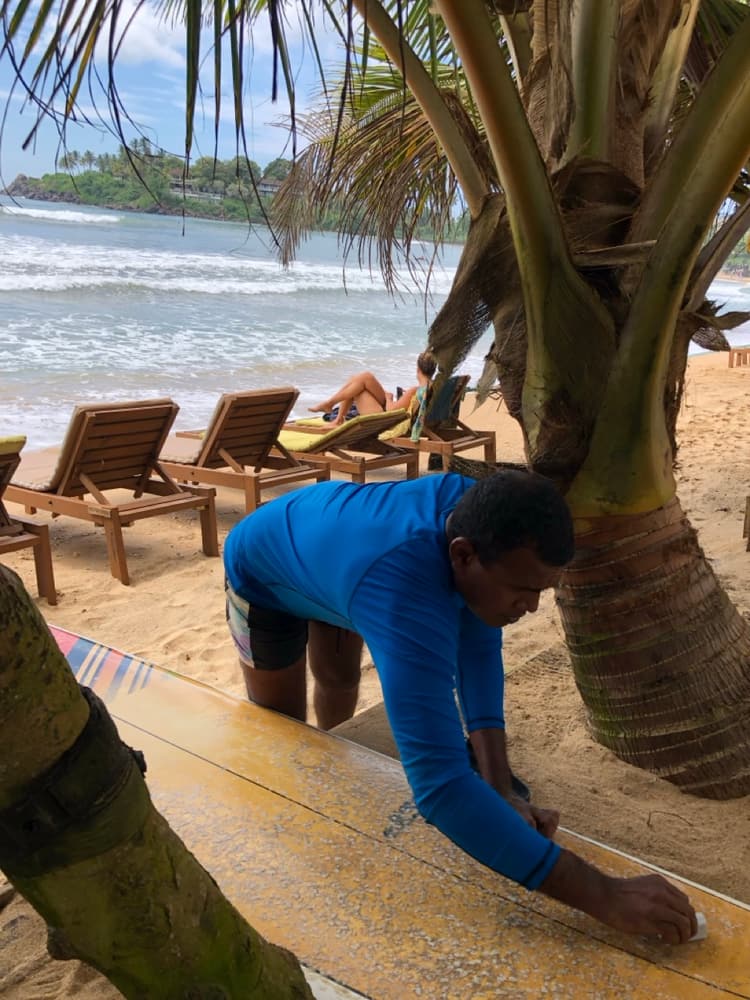 A surf instructor in a blue rash vest waxing a longboard under coconut palms on a south-coast beach