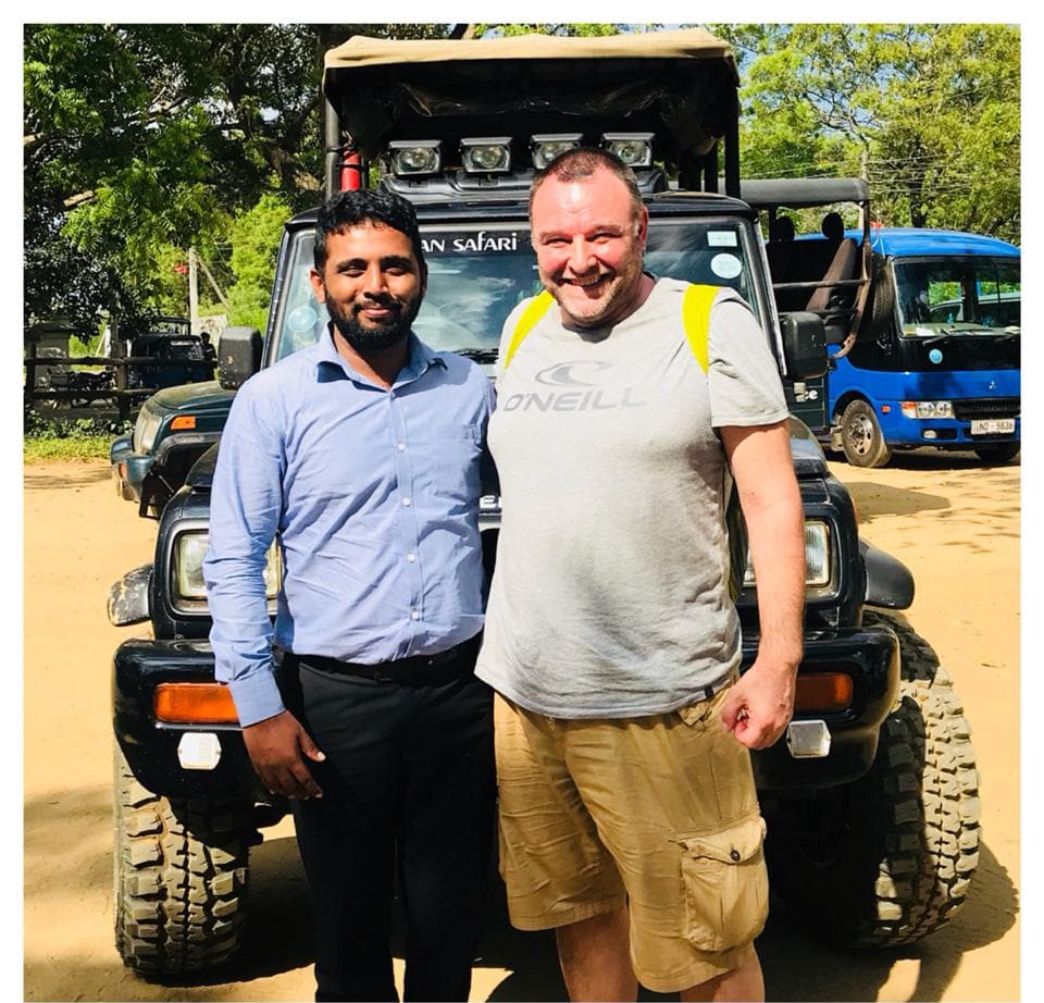 A safari driver-guide in a blue shirt standing beside a guest in front of a safari jeep