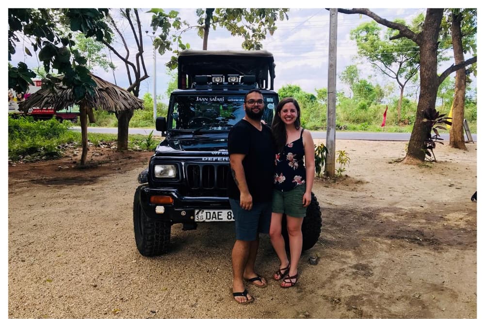 Two guests standing together in front of a black safari jeep parked under trees