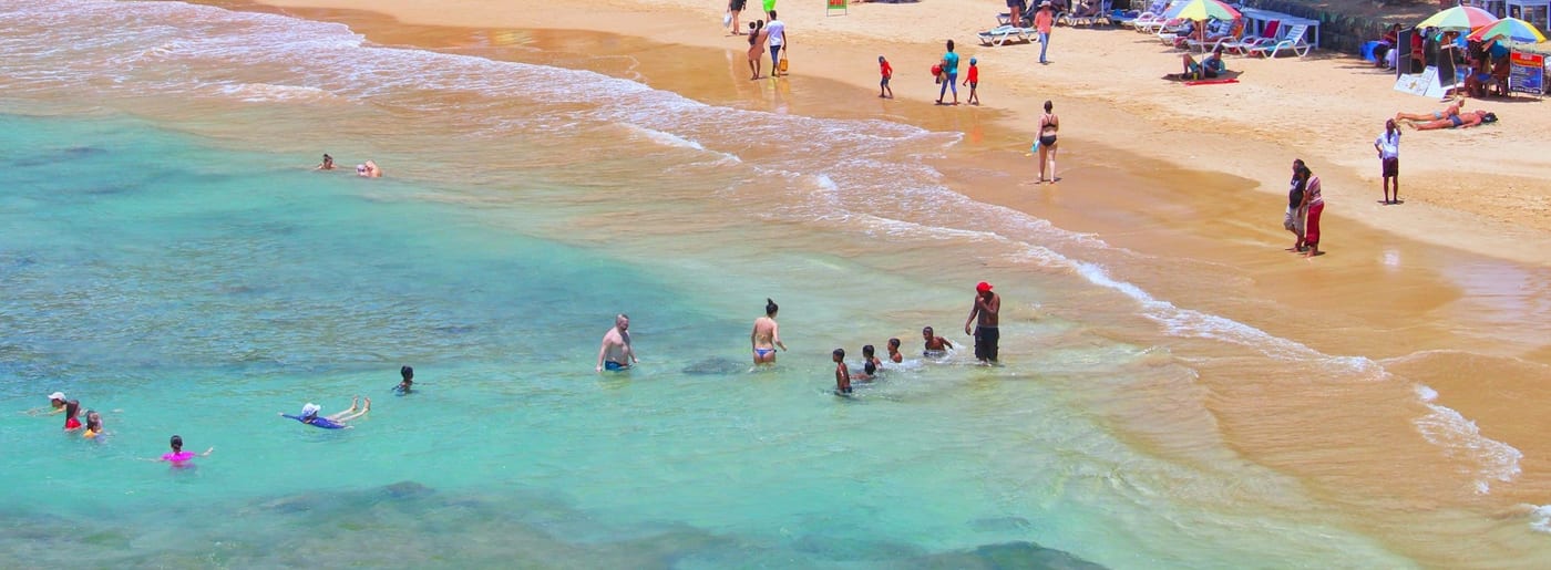 Swimmers in clear turquoise shallows along a curving sandy beach on the south coast