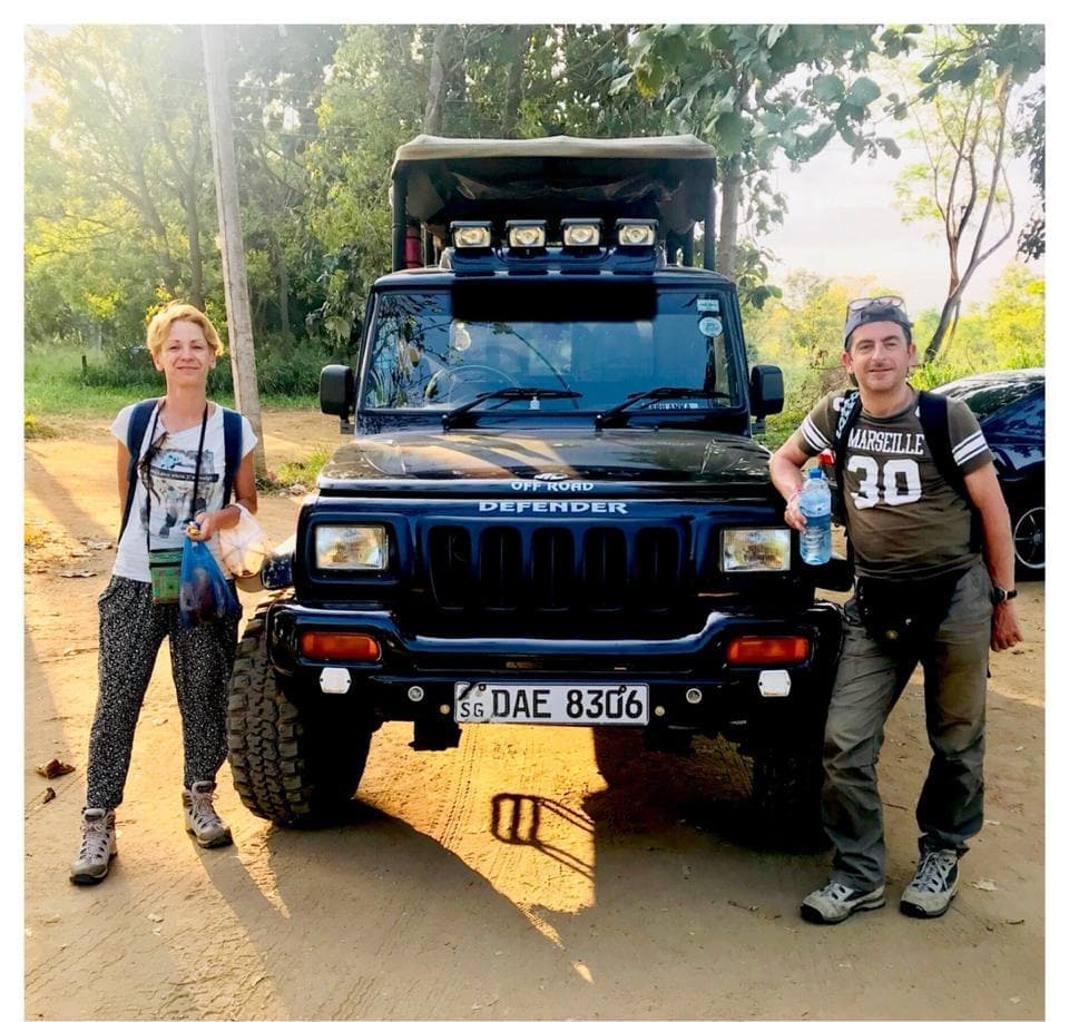Two guests standing either side of a black 4WD safari jeep on a dirt track in low golden light